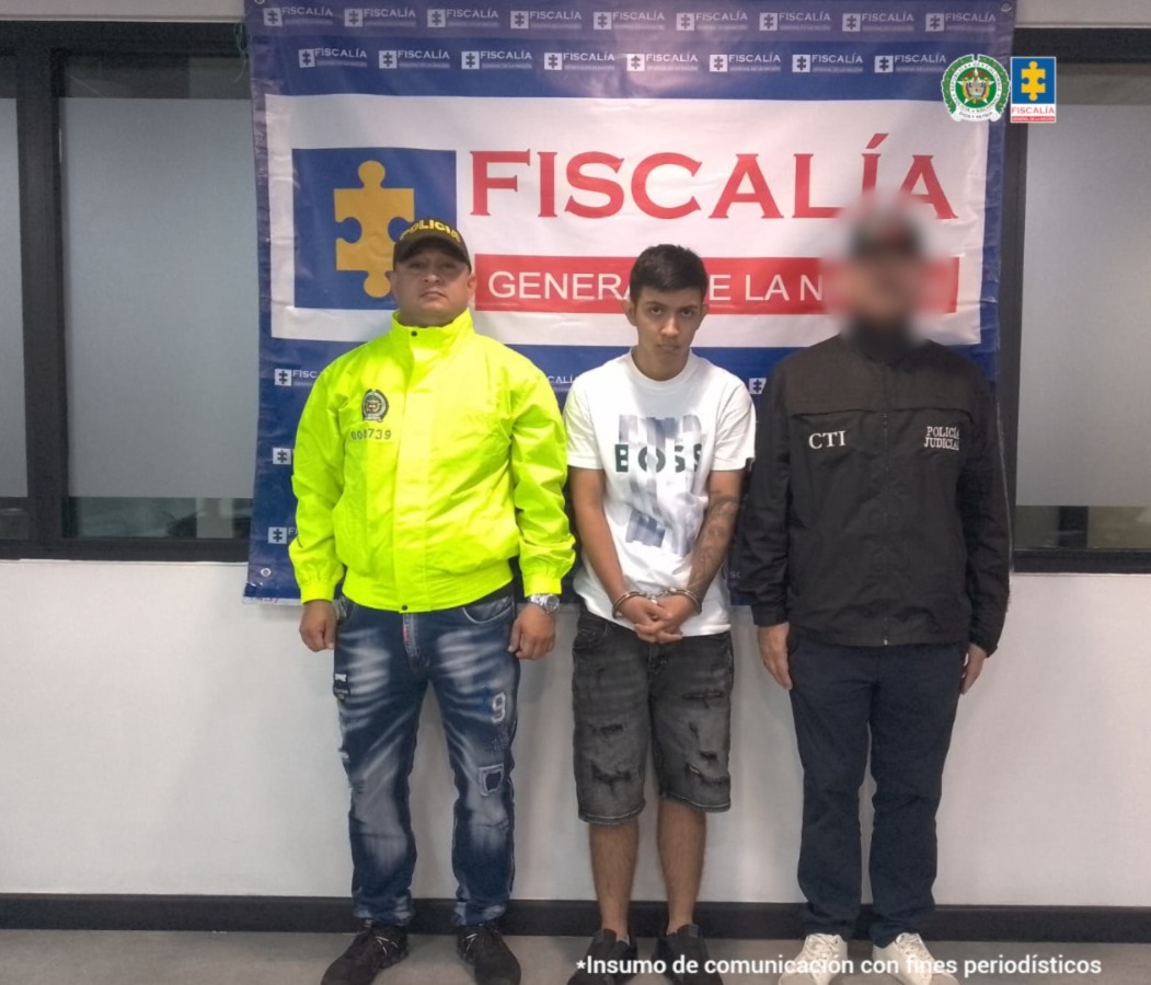 Se visualiza al capturado junto a un uniformado de la Policía Nacional e investigador del CTI. Detrás de ellos el banner que identifica a la Fiscalía General de la Nación.