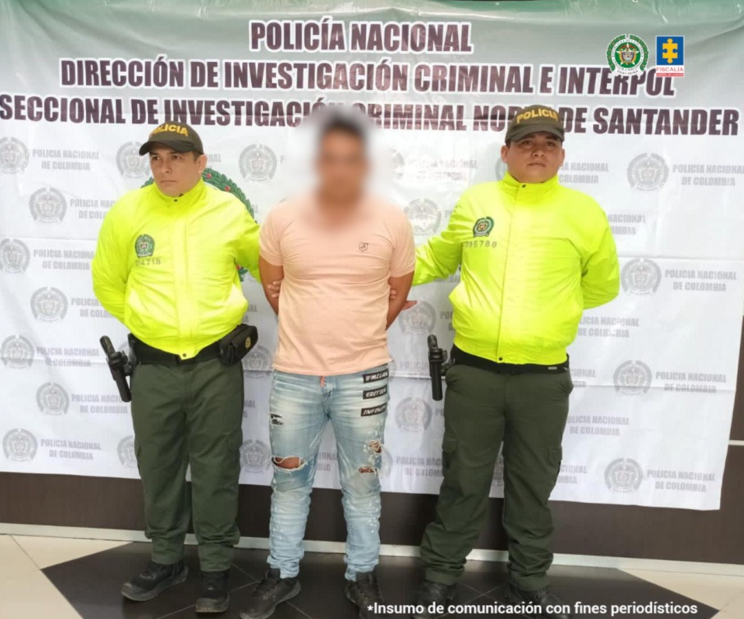 Se visualiza al capturado junto a dos uniformados de la Policía Nacional. Detrás el banner institucional que identifica a la Dirección de Investigación de la Policía Nacional en Norte de Santander