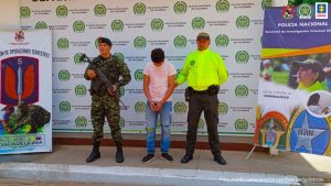 capturado de frente manos esposadas adelante custodiado por un policía y un militar. Detrás banner de Policía y Ejército Nacional.