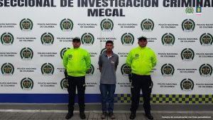 En la fotografía se aprecia de pie y con los brazos atrás a Jaider Yesid Estrella Vele. Viste camiseta de manga corta de color gris, jean azul oscuro y zapatos cafés con nego. A sus extremos hay uniformados de la Policía Nacional. Detrás de ellos hay un pendón institucional de la Policía.