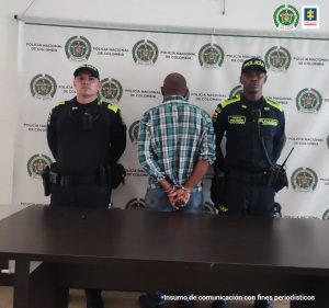 En la fotografía se aprecia a Jairo Cortez Madrid, de pie y de espaldas a la cámara. Viste camisa de manga larga a cuadros, de colores, verde y gris; jean azul. A lado y lado de él hay uniformados de la Policía Nacional. En la parte posterior hay un pendón institucional de la Policía.