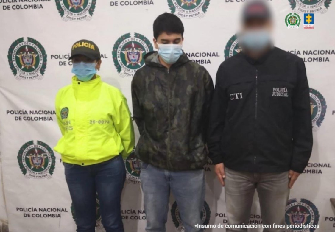 Se observa al hoy condenado en el momento de la captura. A un costado lo custodia un funcionario del CTI y al otro , uno de Policía. Al fondo un pendón de Policía