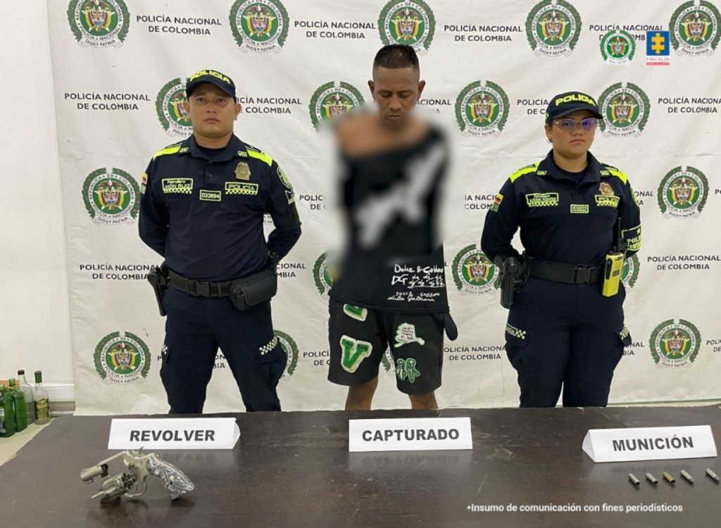 En la imagen aparece un hombre de camiseta negra y pantaloneta negra, entre dos uniformados de la Policía Nacional y frente a una mesa con un arma de fuego y munición.