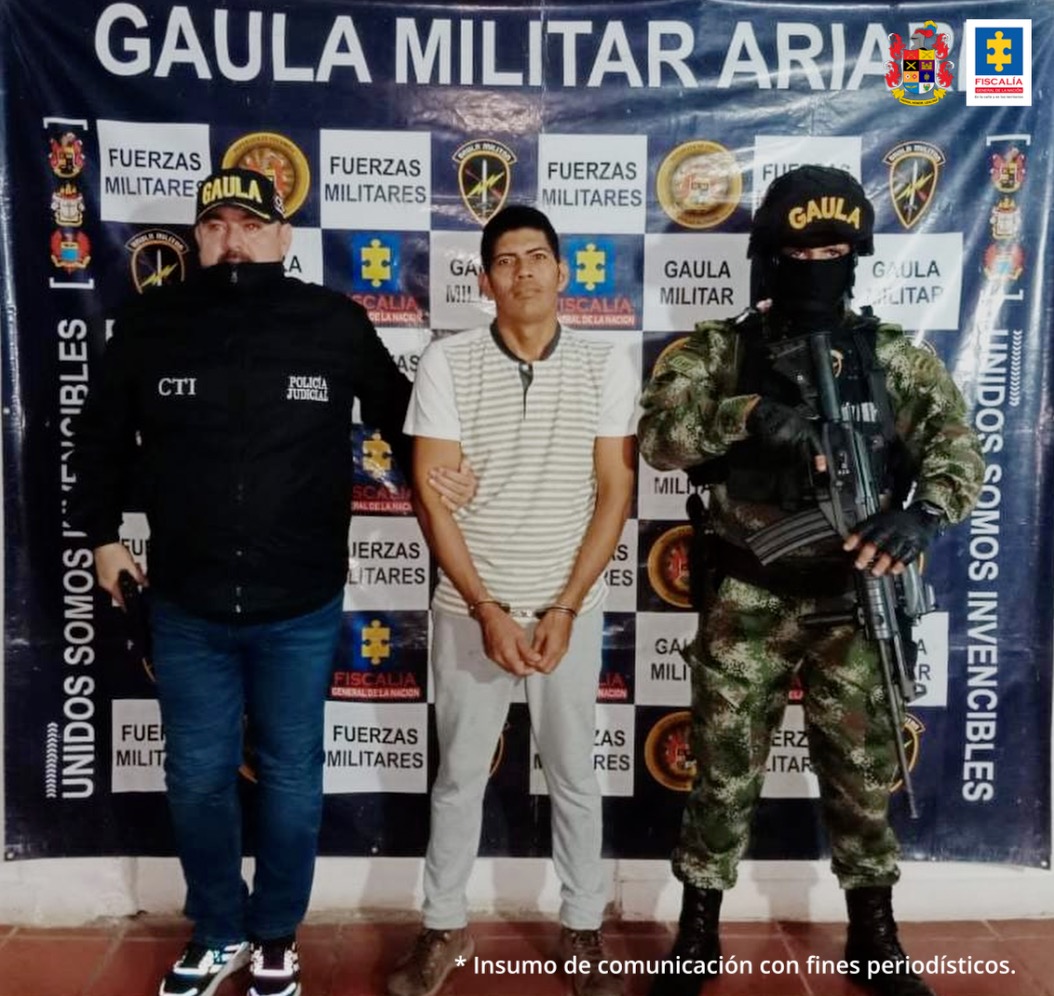 capturado esposado manos al frente custodiado por investigador CTI y militar del Ejército Nacional. Detrás banner del Guala Militar Ariari.