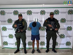 El procesado se encuentra esposado y en custodia por uniformados del Gaula de la Policía Nacional. Detrás suyo un backing de la Policía.