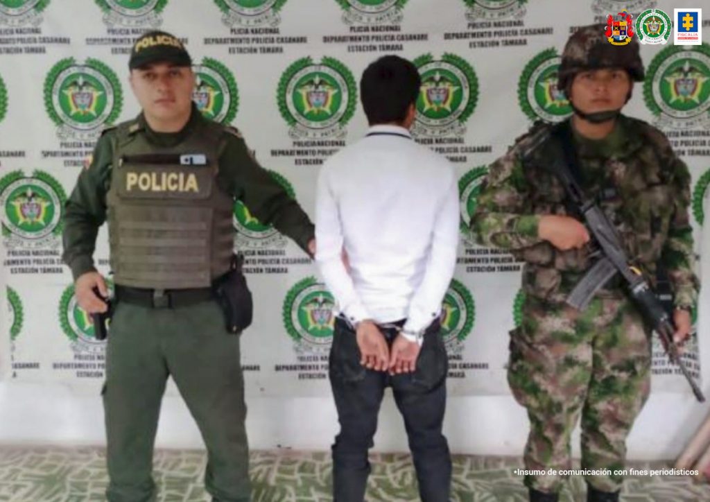 Capturado de espaldas esposado, custodiado por dos uniformados de la Policía Nacional. Detrás banner de la Policía Nacional.