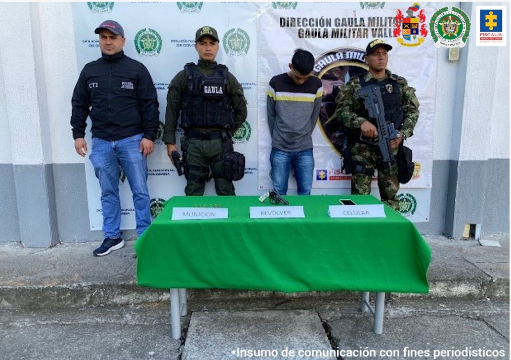 En la foto aparece James Smith Cardona, alias ‘James’ o ‘El Brujo’, de pie, con los brazos atrás y la cabeza agachada. Viste buzo gris con franja amarilla y negra en la parte superior, jean azul desteñido. A su derecha aparece un servidor del CTI de la Fiscalía y un uniformado del Gaula Policía y a la izquierda un uniformado del Gaula Militar. Delante de ellos hay una mesa rectangular con mantel verde y sobre ella hay un revólver, munición y un teléfono celular con letreros que así lo indican. Detrás de ellos hay dos pendones institucionales de la Policía y el Gaula Militar.