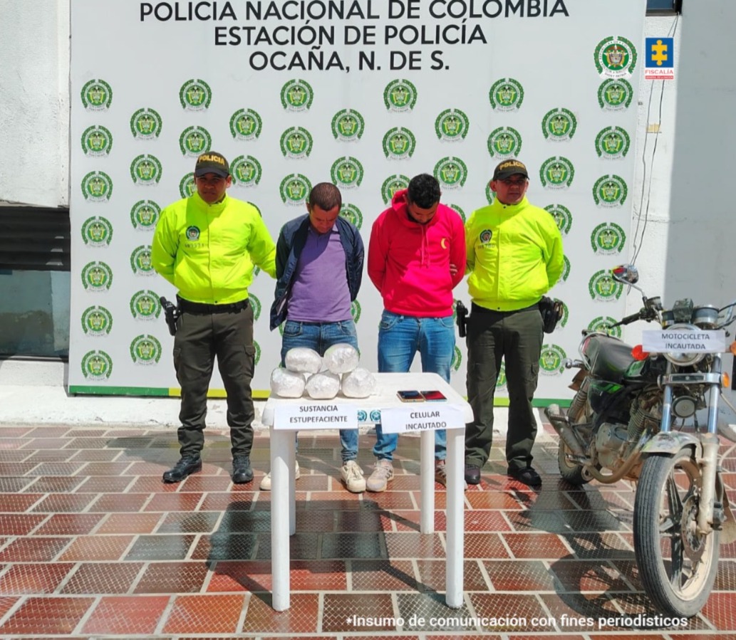 Se visualiza a los capturados junto a dos uniformados de la Policía Nacional. Frente a ellos los elementos incautados como la sustancia estupefaciente, los teléfonos celulares y la motocicleta en que se desplazaban. Detrás de ellos el banner que identifica a la Policía Nacional.