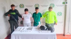 En la fotografía se aprecia de pie y con los brazos atrás a Jean Carlos López Muñoz y Damaris Hurtado Ramírez. La mujer viste camiseta de manga corta blanca con estampado a la altura del pecho y pantalón gris. El hombre viste camiseta de manga corta color verde y pantalón azul. Al lado y lado de ellos hay uniformados de la Policía Nacional. Delante de ellos hay una mesa con mantel blanco y sobre ella hay tres bolsas plásticas negras, una transparente, una gramera y bolsas plásticas. En la parte de atrás de ellos hay un pendón institucional de la Policía Nacional.