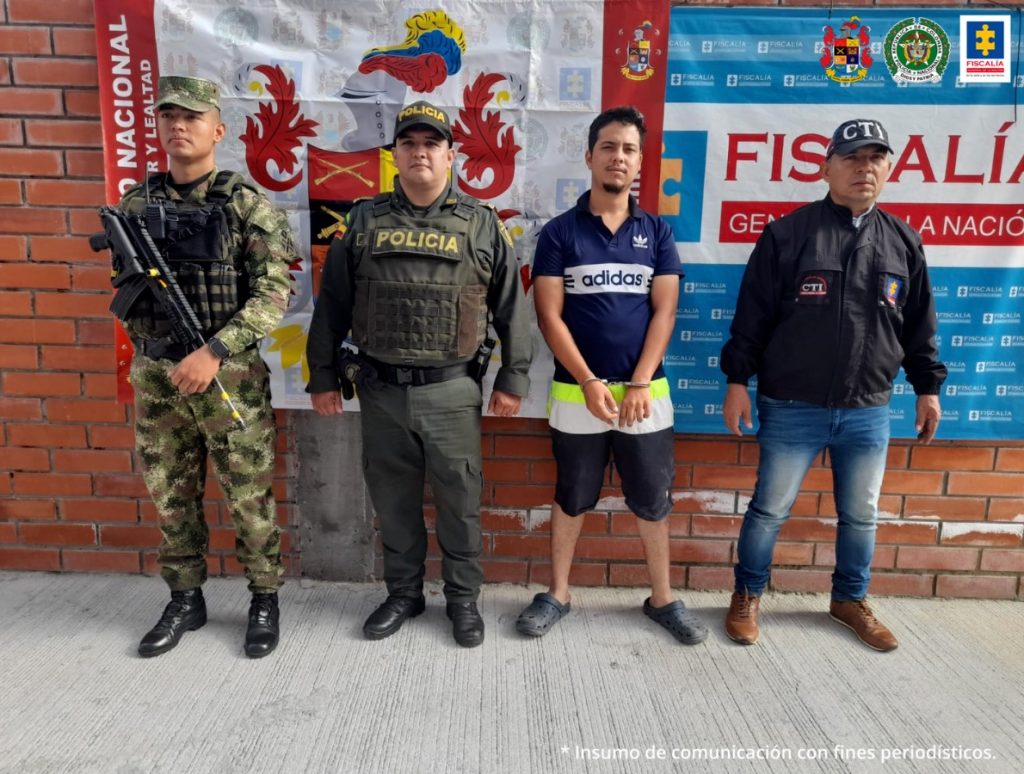 En la fotografía aparece un capturado, junto a personal de Ejército Nacional, Policía Nacional y Fiscalía General de la Nación. En la parte posterior de la imagen se ven banners de Ejército Nacional, Policía Nacional y la Fiscalía General de la Nación.