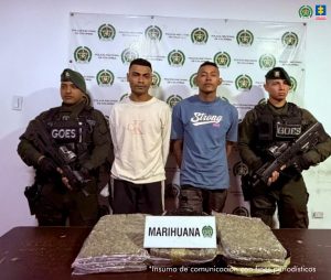 En la fotografía aparecen de pie y con los brazos atrás ⁠Jeiron Antonio Rodríguez Donado y Yesid Ramírez Patiño. Uno, viste con camiseta de color azul, mangas cortas y estampado blanco en el pecho. La otra persona viste con buso de color beage, de mangas largas y pantalón de color negro. En los extremos hay uniformados de la Policía Nacional. Delante de ellos hay una superficie de color marrón y sobre ella se van paquetes grandes de bolsas plásticas transparentes con contenido vegetal. Detrás de ellos hay un pendón institucional de la Policía.
