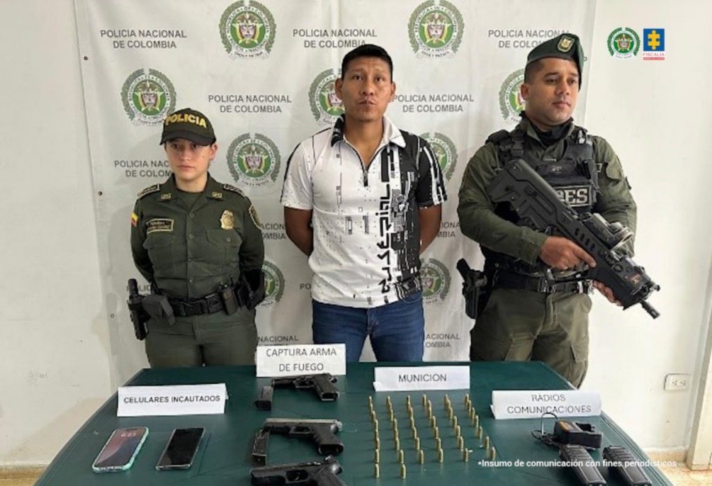 En la fotografía aparece de pie y con los brazos atrás Jeison Eduardo Mesa Ipía. Viste con camisa con cuello, mangas cortas, de color lanco y estampado de color negro en el lado izquierdo; asimismo, jean azul. En sus extremos hay uniformados de la Policía Nacional. Delante de ellos hay una superficie de color verde y sobre ella se aprecian tres armas de fuego, munición, proveedores y celulares. Detrás de ellos hay un pendón institucional de la Policía.