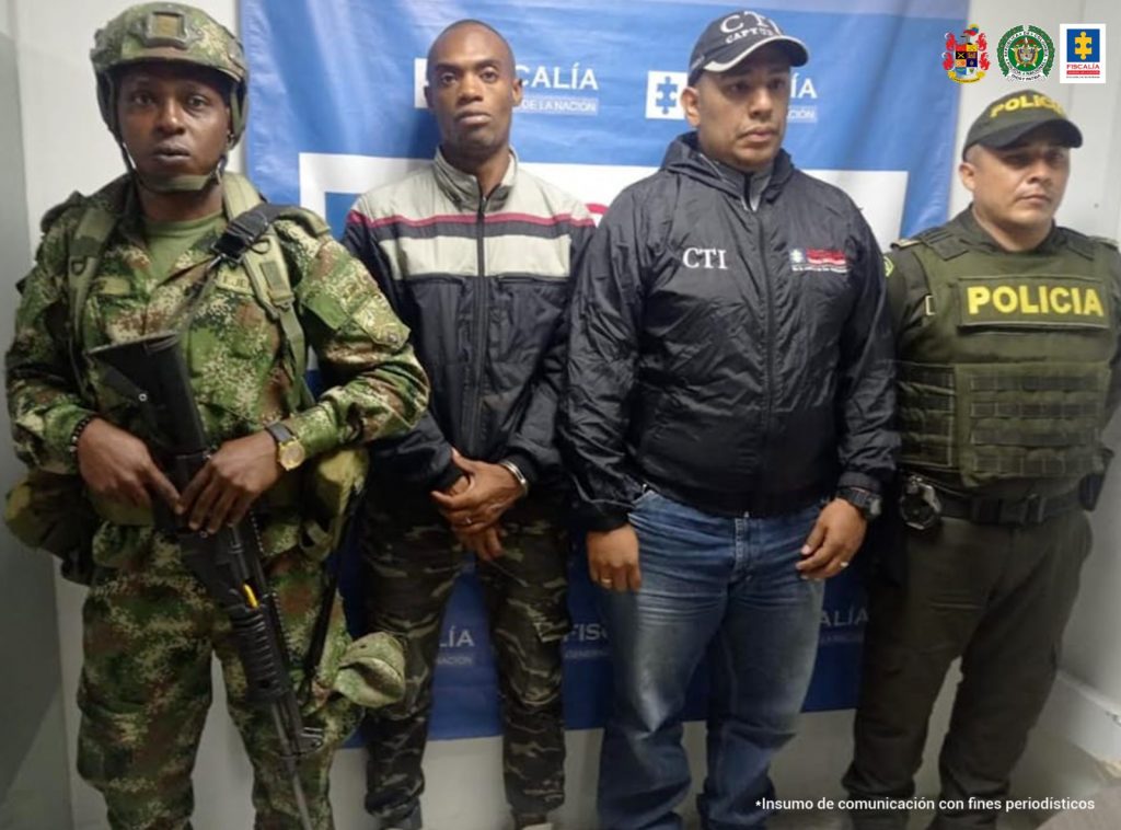 En la fotografía se aprecia a Jeison Stiven Angulo, de pie y con los brazos adelante y esposado. Viste chaqueta azul con franjas grises anchas y franjas delgadas azul y roja en la parte superior; pantalón camuflado y zapatillas blancas. Al lado izquierdo hay un servidor del CTI de la Fiscalía y un uniformado de la Policía Nacional; al lado izquierdo hay un uniformado del Ejército Nacional. Detrás de ellos hay un pendón institucional de la Fiscalía.
