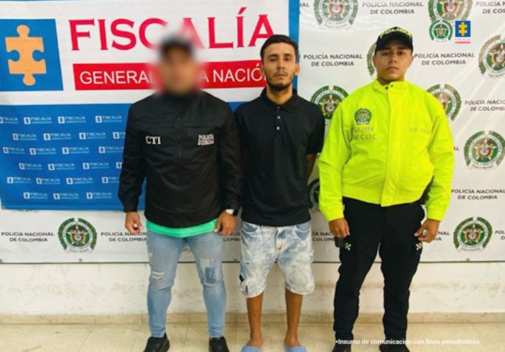 Se visualiza al capturado junto a un investigador del CTI y un uniformado de la Policía Nacional. Detrás de ellos los banner que identifican a la Fiscalía General de la Nación y a la Policía Nacional.