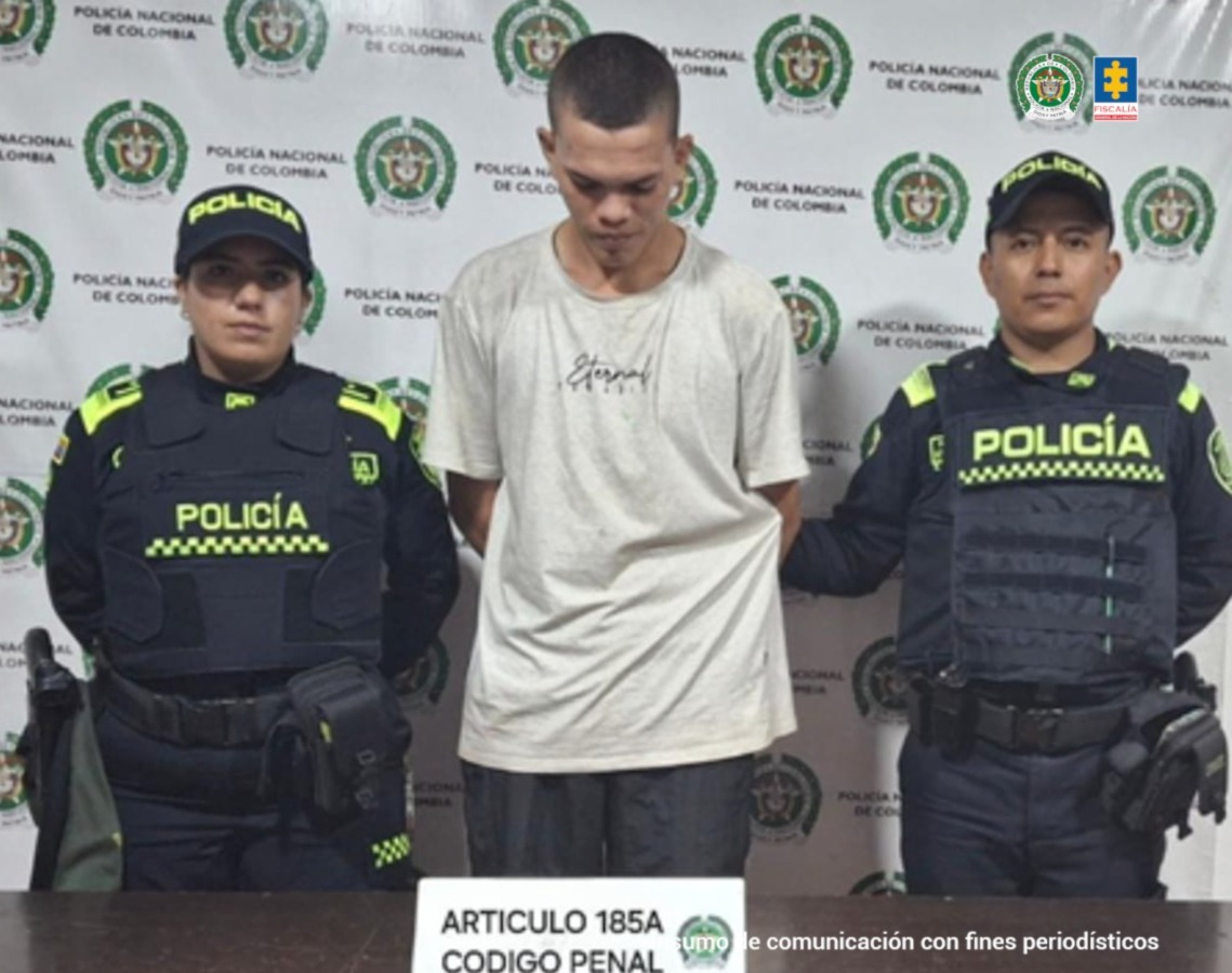 En la fotografía se aprecia de pie y con los brazos atrás a Jerson Esneyder Gutiérrez Giraldo. Viste camiseta de color blanco, de manga corta y pantalón oscuro. A sus extremos hay uniformados de la Policía Nacional. Delante de ellos hay una superficie de color marrón y sobre ella hay un letrero que dice: ‘Artículo 185A Código Penal’. Detrás de ellos hay un pendón institucional de la Policía.