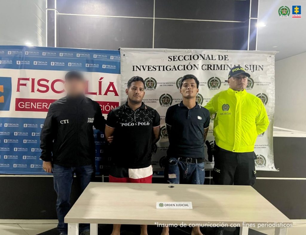 Se observa a los dos capturados junto a un investigador del CTI y un uniformado de la Policía Nacional. Detrás de ellos los banners de la Fiscalía General de la Nación y de la Seccional de Investigación Criminal Sijín de la Policía Nacional.