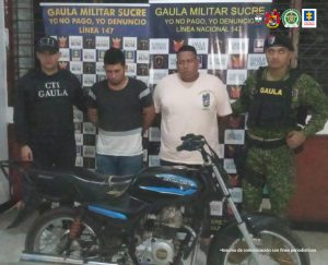 En la imagen se ven dos personas detenidas, frente a una motocicleta y entre dos uniformados de la Fiscalía y el Gaula.