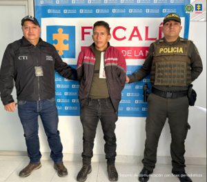 En la fotografía se aprecia de pie y con los brazos atrás a Jesús Bernardo Mamián. Viste con camiseta de color verde oscuro y sobre ella una chaquela que en la parte superior es de color vinotinto y en la parte de abajo verde oscuro; asimismo, pantalón y zapatos negros. A su derecha hay un servidor del CTI de la Fiscalía. A su izquierda un uniformado de la Policía Nacional. Detrás de ellos hay un pendón institucional de la Fiscalía.