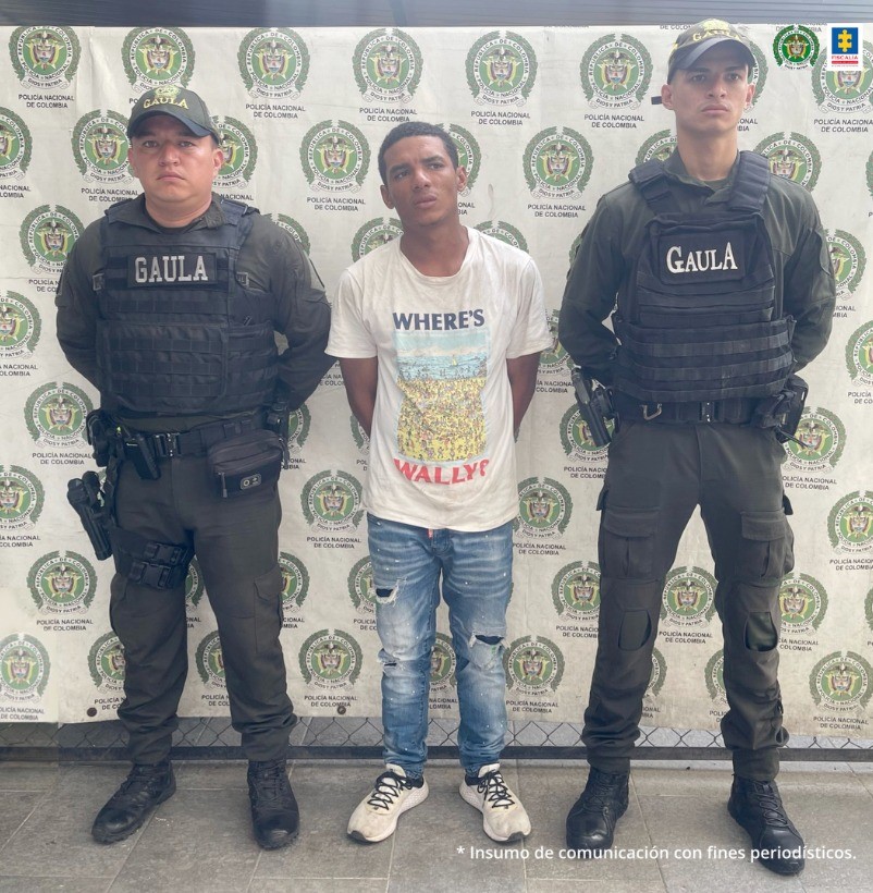 capturado de frente custodiado por dos policías. Detrás banner de la Policía Nacional.