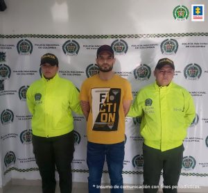 En la fotografía se aprecia de pie y con los brazos atrás a Jesús Guillermo Ortiz Delgado. Viste con gorra oscura, camiseta de manga corta color mostaza con estampado negro, blanco y amarillo; jea azul oscuro. A lado y lado de él hay uniformados de la Policía Nacional. Detrás de ellos hay un pendón institucional de la Policía.