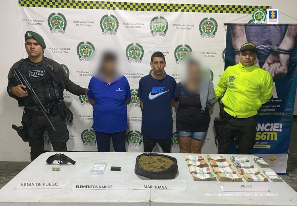 En la fotografía se aprecia de pie, con los brazos atrás Jesús Hernán Gómez, alias ‘Lápiz’. Viste camisa de manga corta azul oscuro con la palabra ‘Nike’ y el logo en blanco; asimismo, pantalón negro. A lado y lado de él hay un uniformado de la Policía Nacional. Delante de ellos hay una superficie blanca y sobre ella hay dinero en efectivo, una bolsa negra con contenido vegetal y otros elementos. Detrás de ellos hay un pendón institucional de la Policía.