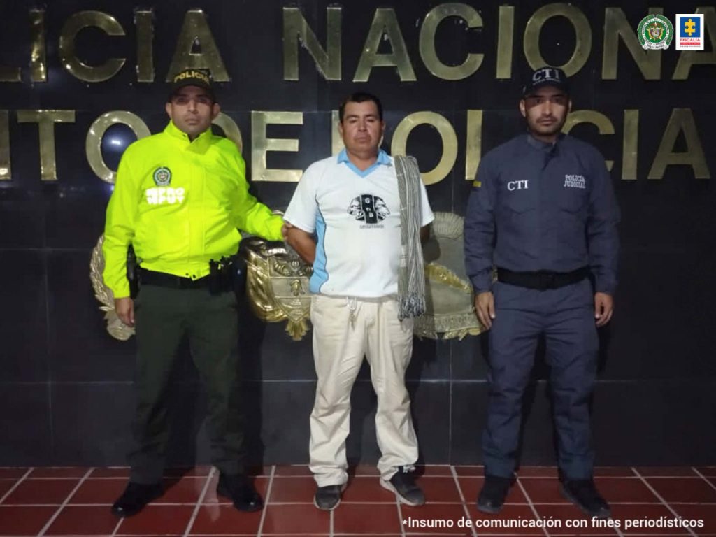 En la fotografía aparecen dos capturados, junto a personal de Fiscalía General de La Nación y Policía Nacional. En la parte posterior de la imagen se ven banners de Policía Nacional y la Fiscalía General de la Nación.    