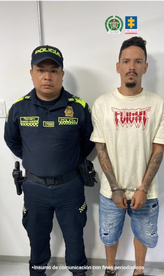 En la fotografía aparece de pie, con los brazos adelante y esposado Jesús Orlando Holguín Velasco. Usa barba tipo ‘chivera’; viste con camiseta de manga corta, de color beage y con estampado rojo a la altura del pecho; asimismo, usa jean azul claro desgastado. A su extremo derecho hay un uniformado de la Policía Nacional.