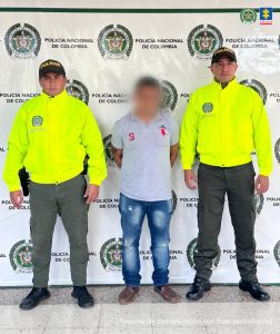 El procesado se encuentra custodiado por dos uniformados de la Policía Nacional. Detrás se observa un backing de la Policía. El agresor tiene cubierto el rostro.