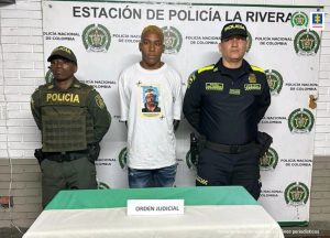 En la foto se aprecia de pie a Jetli David Quijano. Viste camiseta blanca con estampado multicolor del rostro de un hombre, a la altura del pecho; asimismo, jean azul. A lado y lado de ella hay uniformados de la Policía Nacional. Delante de ellos hay una mesa con mantel de colores blanco y verde y sobre ella hay un letrero que dice: ‘Orden judicial’. Detrás de ellos hay un pendón institucional de la Policía.