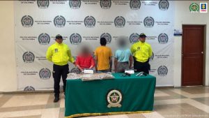 Se observan tres personas capturadas de espaldas. Las dos de los costados con un efecto de difuminado ya que no fueron judicializadas. A cada costado funcionarios de Policía nacional. Atrás un pendón de Policía Nacional