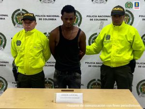 En la fotografía se aprecia a Jhoan Stiward Sánchez Bazán, alias Zapato, de pie y con los brazos atrás. Viste camiseta tipo esqueleto de color negra, pantalón y zapatos negros. A lado y lado de él hay dos uniformados de la Policía Nacional. Delante de ellos hay una mesa y sobre ella un letreo que dice: ‘Orden judicial concierto para delinquir’. Detrás de ellos hay un pendón de la Policía.
