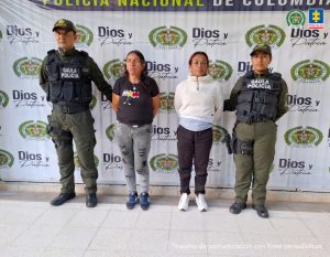 Jhoana Paola Riaño Neuta y Seleny Facundo Toledo capturadas en Bogotá fueron imputadas po rla Fiscalía por extorsión. Texto Alternativo: capturadas de frente custodiadas por dos policías. Detrás banner de la Policía Nacional.