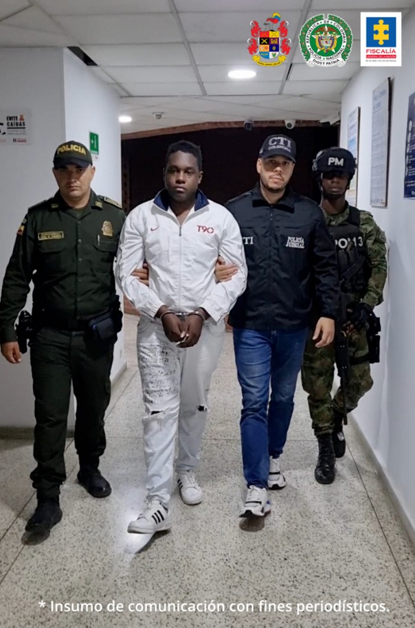 En la fotografía se aprecia, en acción de caminata y esposado a Jhoason Estiven Camacho Mosquera. Viste camisa de manga larga blanca; pantalón y tenis blancos. Al lado izquierdo hay un servidor del CTI de la Fiscalía y un uniformado del Ejército. Al lado derecho hay un uniformado de la Policía Nacional.