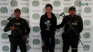 En la imagen está el presunto responsable del homicidio de la profesora con dos servidores de la Policía Nacional a los lados, y atrás está el pendón con el logo de esa institución