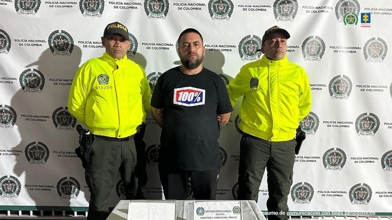 El capturado aparece en el centro de la imagen, rodeado de dos miembros de la Policía. Tiene las manos esposadas en la espalda y detrás de él hay un backing de la Policía.
