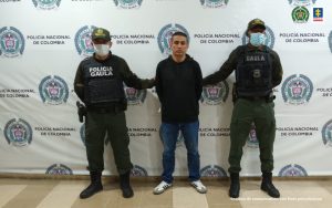 Se observa en el centro el capturado, a cada costado un funcionario de Policía Nacional y al fondo un pendón de Policía Nacional
