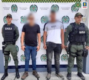 En la foto aparecen, de pie Jhon Alexánder Zapata Varón y Ánderson Muñoz Mosquera. Uno de ellos viste camiseta negra de manga corta y jean azul. La otra persona viste camiseta blanca y gris, de manga corta; así como jean oscuro. Al lado y lado de ellos hay uniformados del Gaula de la Policía Nacional. Detrás de ellos hay un pendón institucional de la Policía.