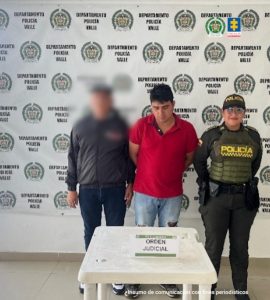 En la fotografía se aprecia de pie y con los brazos atrás a Jhon Anderson Peñaloza Urrea. Viste con camisa de mangas cortas y cuello, de color rojo; así como jean azul claro desgastado. En su extremo derecho hay un servidor del CTI de la Fiscalía y en el derecho una uniformada de la Policía Nacional. Delante de ellos hay una mesa blanca y sobre ella hay un letrero que dice: ‘Orden judicial’. Detrás de ellos hay un pendón institucional de la Policía.