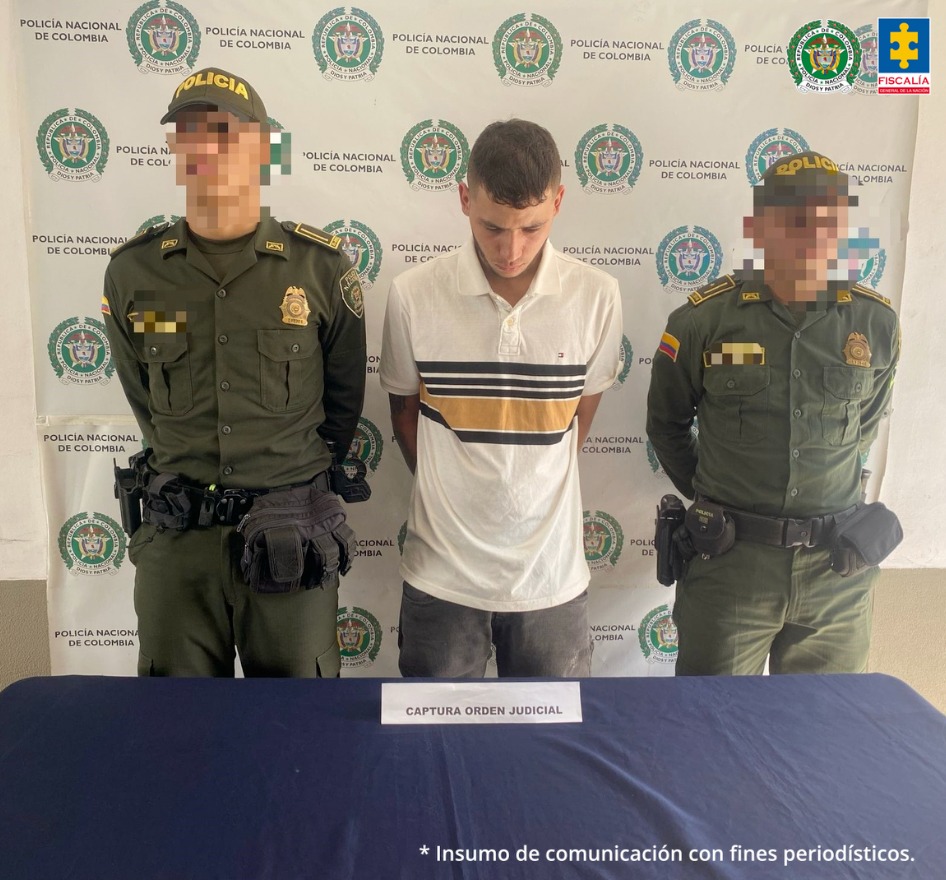 En la fotografía se aprecia de pie y con los brazos atrás a Jhon Bairon Largo Ramírez, alias Bagre. Viste camisa de amnga corta color beage con franja amarillo mostaza y líneas negras horizontales a la altura del pecho; pantalón gris. A lado y lado de él hay uniformados de la Policía Nacional. Delante de ellos hay una superficie azul y sobre ella un letrero que dice: ‘Captura orden judicial’. Detrás de ellos hay un pendón institucional de la Policía.