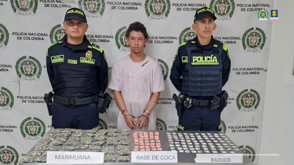 En la fotografía se aprecia de pie, con los brazos adelante y esposado a Jhon David Torres Ángel. Viste camiseta de color palo de rosa con estampado blanco en forma de visto bueno la altura del pecho. A sus extremos hay uniformados de la Policía Nacional. Delante de ellos hay una superficie gris y sobre ella hay gran cantidad de bolsas plásticas, pequeñas y transparentes con contenido blanco, rosado y verde. Asimismo, tres letreros que dicen: ‘Marihuana’, ‘Base de coca’, ‘Bazuco’. Detrás de ellos hay un pendón institucional de la Policía.