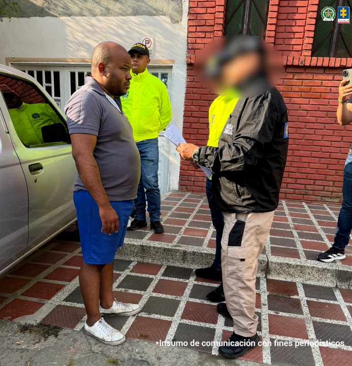 La fotografía fue tomada en exteriores. Aparece de pie Jhon Edinson Mina Gutiérrez. Viste con camisa de cuello y manga corta, de color gris; bermuda de color azul rey y zapatillas blancas. Frente a él hay un servidor del CTI de la Fiscalía y un uniformado de la Policía Nacional. En el extremo izquierdo del judicializado hay otro uniformado de la Policía.
