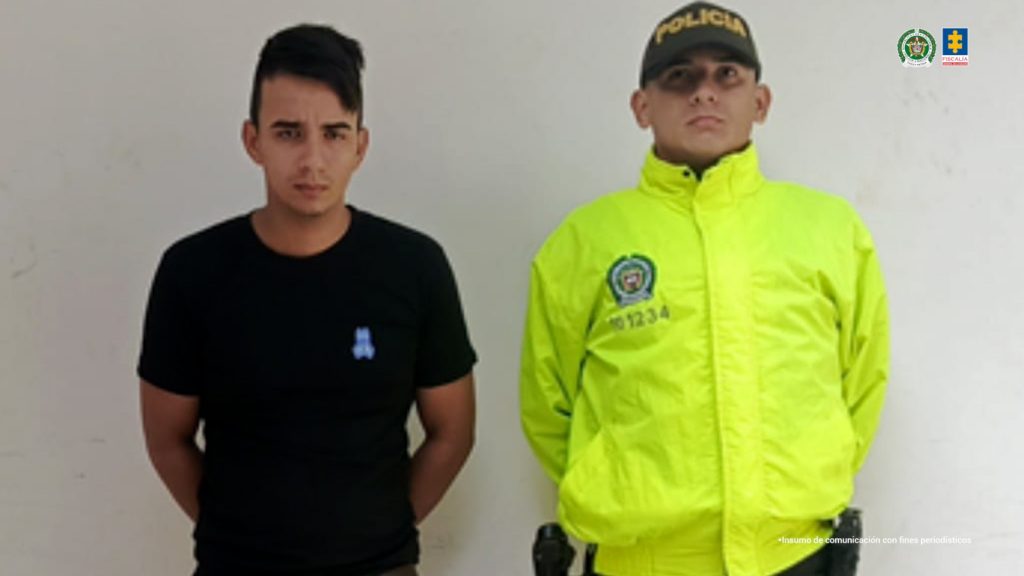 En la fotografía se aprecia de pie y con los brazos atrás a Jhon Eduar Foronda Euse, alias Jhon. Viste con camiseta de manga corta de color negro, jean azul desgastado y pantuflas de color gris con blanco. A su extremo izquierdo hay un uniformado de la Policía Nacional.