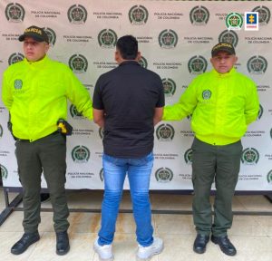  En la fotografía aparece un capturado, junto a personal de Fiscalía General de la Nación y Policía Nacional. En la parte posterior de la imagen se ven banners de Policía Nacional y la Fiscalía General de la Nación.     