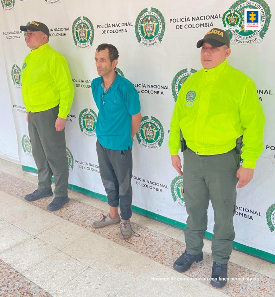 En la fotografía aparece un capturado, junto a personal de Policía Nacional. En la parte posterior de la imagen se ven banners de Policía Nacional y la Fiscalía General de la Nación.  