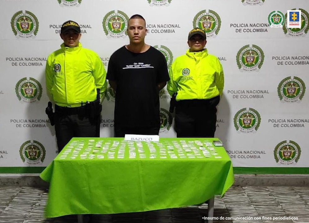 En la foto aparece de pie Jhon Hader Osorio Pizarro con los brazos atrás; vestido con camiseta negra y pantalón oscuro. A lado y lado de él hay dos uniformados de la Policía Nacional. Delante de ellos hay una mesa con mantel verde y sobre ella gran cantidad de bolsas plásticas y un letrero que dice ‘Bazuco’. Detrás de ellos hay un pendón institucional de la Policía.