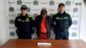 Se observa un hombre de frente y a cada costado un uniformado de Policía Nacional. Atrás un pendón de Policía Nacional.  Al frente una mesa con el aviso ACTO SEXUAL