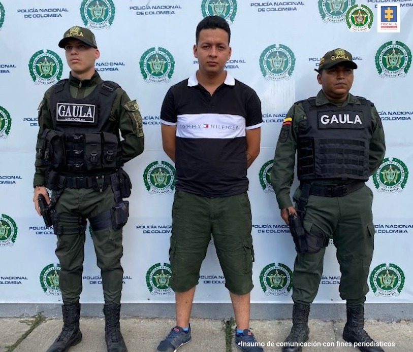 En la foto aparece Jhon Jaber Salinas Serna, con los brazos atrás; vestido con camiseta negra con cuello blanco y una franja blanca a la altura del pecho; asimismo, bermuda verde y tenis oscuros. A lado y lado de él hay dos uniformados de la Policía Nacional con chalecos que tienen la palabra ‘Gaula’. Detrás de ellos hay un pendón institucional de la Policía Nacional.