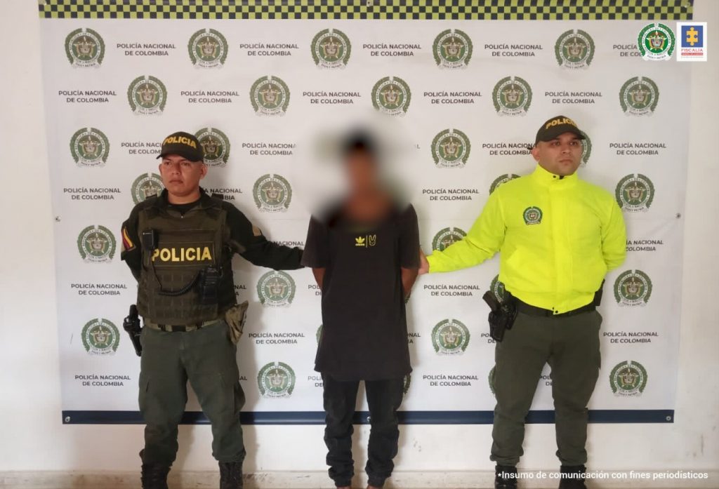 En la imagen está el procesado, con el rostro distorsionado y dos servidores de la Policía Nacional a los lados, con el pendón de esa entidad en la parte de atrás