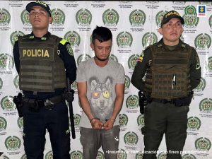 capturado de frente esposado manos atrás, custodiado por dos policías. Detrás banner de la Policía Nacional.