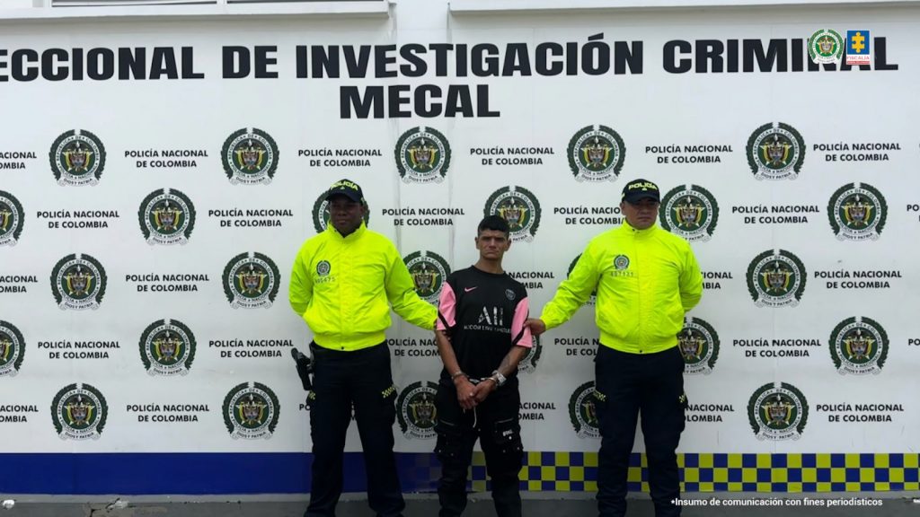 En la fotografía se aprecia de pie, con los brazos adelante y esposado a Jhon Jairo Cardona Sánchez. Viste camisa de color negro con estampados en los extremos derecho e izquierdo y en la parte central. Las mangas de la camisa son cortas y de color fuccia, con una franja negra diagonal. Asimismo, usa pantalón negro con varios bolsillos. A sus extremos hay uniformados de la Policía Nacional. Detrás de ellos hay un pendón institucional de la Policía.
